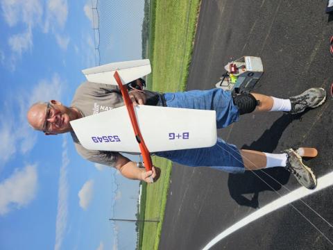 Man holding a model airplane on an asphalt surface, smiling under a clear blue sky.