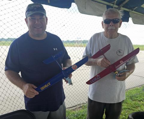 Two men smiling, holding model airplanes, standing outdoors by a wire fence.