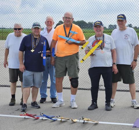 A group of older men holding model planes, standing on a tarmac.