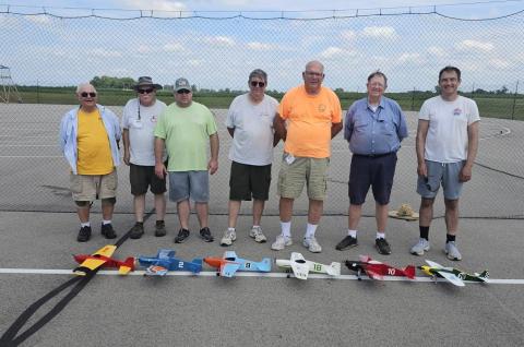 Seven men standing behind model airplanes on a tarmac, under a blue sky.