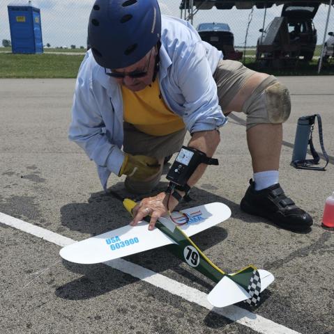 Man in helmet kneeling on pavement, adjusting a small model airplane.