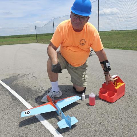 Man kneeling beside a model airplane on a runway, wearing a blue helmet and orange shirt.