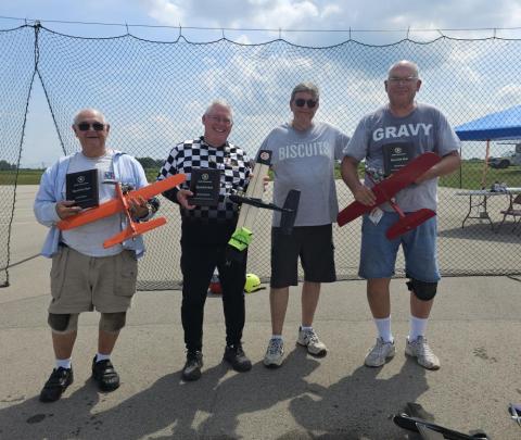 Four men proudly holding model airplanes and awards outdoors under a cloudy sky.