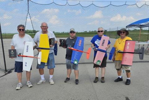 Five men proudly holding colorful model airplanes at an outdoor event.