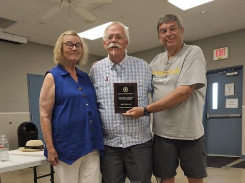Three people smiling, one holding a plaque in a casual indoor setting.