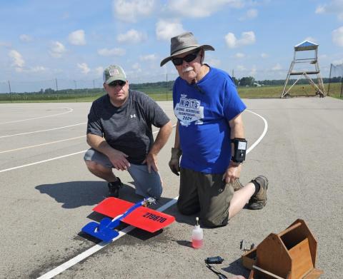 Two men kneeling by a red and blue model plane on a paved runway.