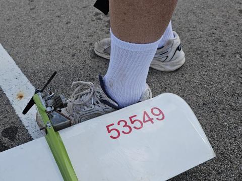 Toy plane wing with "53549" on pavement near sneakered foot.