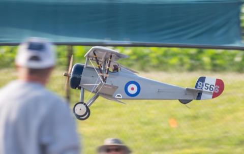 Model biplane with RAF roundel flying in a grassy field, blurred person in foreground.