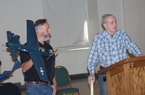 Two men standing; one holds a model navy plane, the other speaks at a podium.