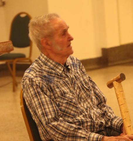 Elderly man sitting on a chair indoors, holding a cane, wearing a plaid shirt.