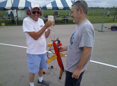 Two men smiling, holding a small red and yellow rocket on a runway.