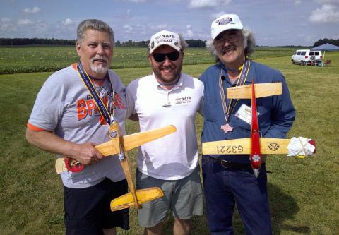 Three men smiling outdoors, each holding a model airplane and wearing medals.