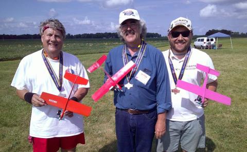 Three men holding colorful model airplanes, standing on grassy field.