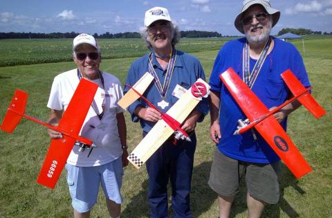 Three people outdoors holding toy airplanes, smiling.