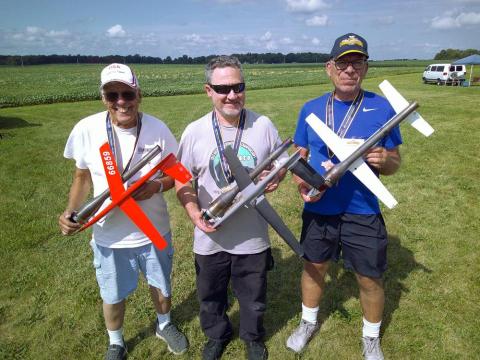 Three men smiling, holding model airplanes on a grassy field under a blue sky.