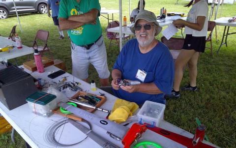 Man sitting at a table with tools, smiling at an outdoor event.