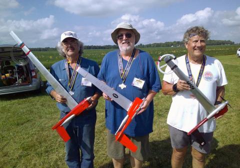 Three smiling people holding model airplanes on a grassy field.