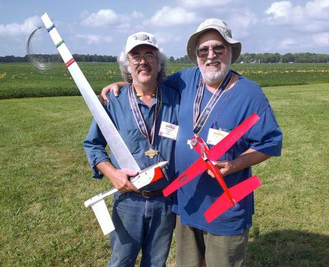 Two men smiling, holding model airplanes in a grassy field.