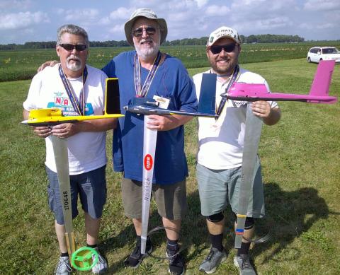 Three men outdoors holding model rockets, smiling in a grassy field.