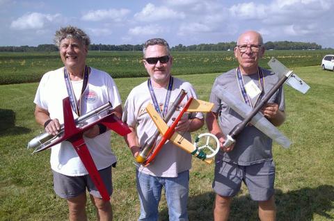 Three people smiling, holding model airplanes, standing in a grassy field.