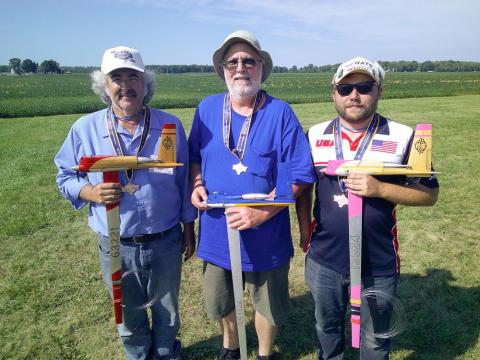 Three men outdoors holding model planes, smiling, on a sunny day with grassy field in background.