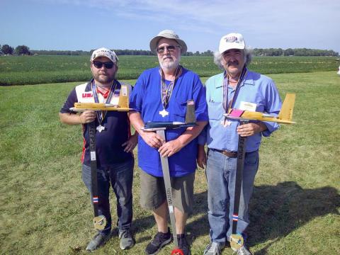 Three people holding small model planes, standing on grass at an outdoor event.