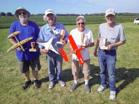 Four people outdoors holding model airplanes and trophies, standing on grass.