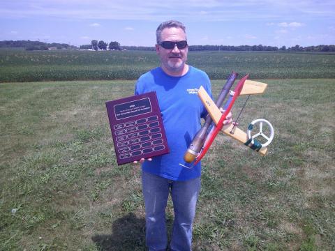 Man holding a model airplane and a plaque in a grassy field.
