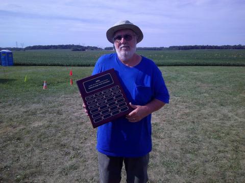 Man in blue shirt holding a plaque on a grassy field, wearing a hat and sunglasses.