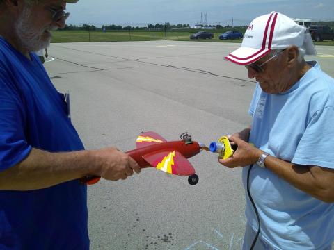 Two men examining a red model airplane on a sunny day.
