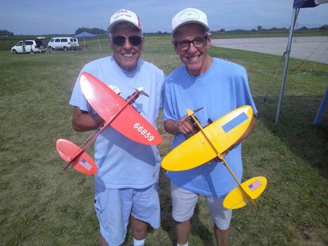 Two smiling men hold colorful model planes outdoors.