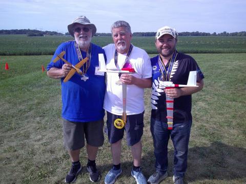 Three men outdoors holding model airplanes, smiling.