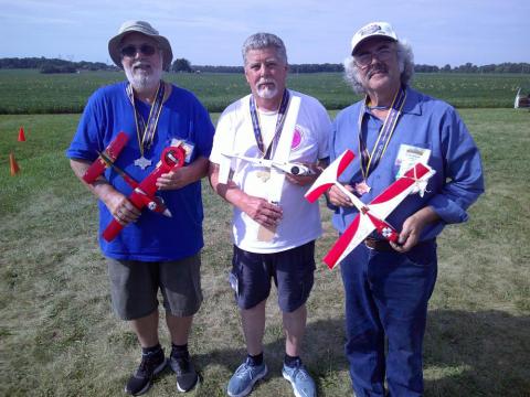 Three men holding model airplanes, standing outdoors with medals.