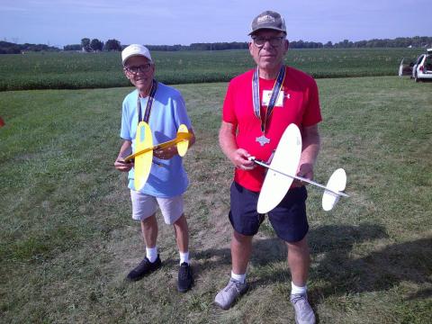 Two men holding colorful model airplanes on a grassy field.
