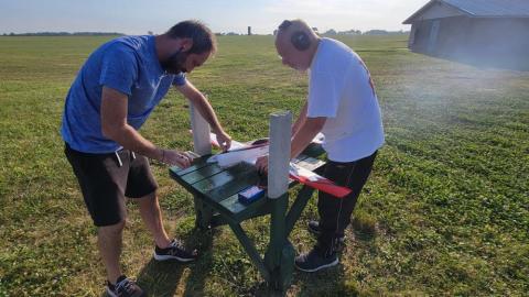 Two men working on a model airplane outdoors on a sunny day.