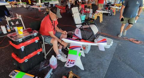 Man in a red shirt working on a model airplane under a canopy.