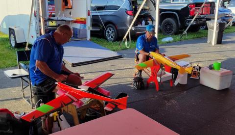 Two people assembling model airplanes under a canopy, surrounded by vehicles.