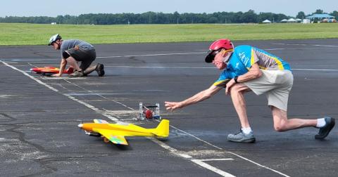 Two people launching model planes on a paved runway, wearing helmets and casual attire.