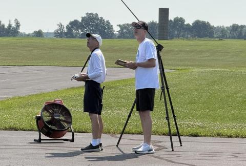 Two people operating remote controls on a paved area near a grassy field, with a large fan nearby.