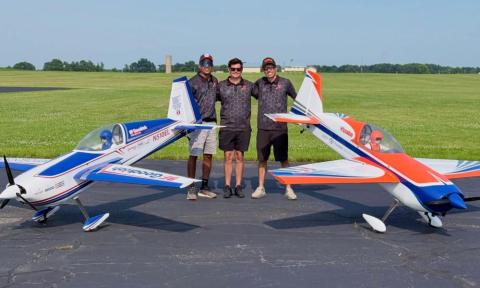 Three people stand between two model airplanes on a tarmac in a grassy field.