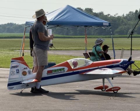Model airplane on pavement with nearby people under a blue canopy.