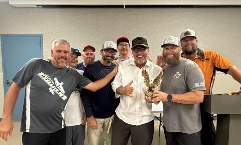 Smiling group of nine men, one holding a trophy, in a casual indoor setting.