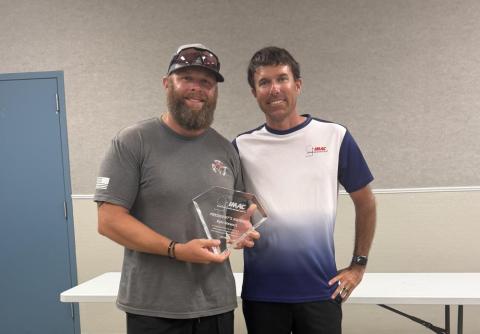 Two men stand smiling indoors, one holding a clear trophy.