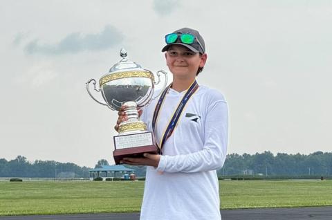 Young person smiling, holding a large trophy outdoors on a cloudy day.