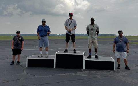 Five people standing on and around podiums on an airfield on a cloudy day.