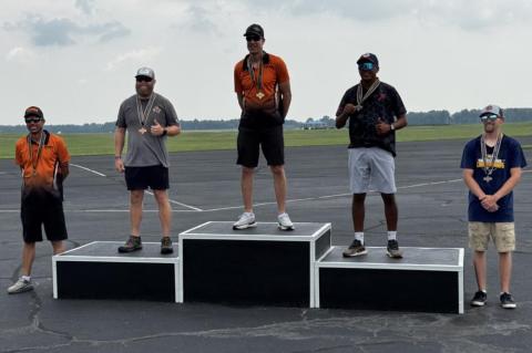 Award ceremony with five men on podiums, each holding medals, outdoors on an airfield.