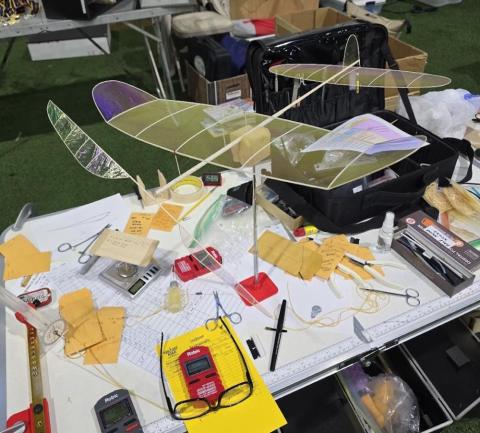 Table with model airplanes, tools, papers, and electronic devices in a workshop setting.