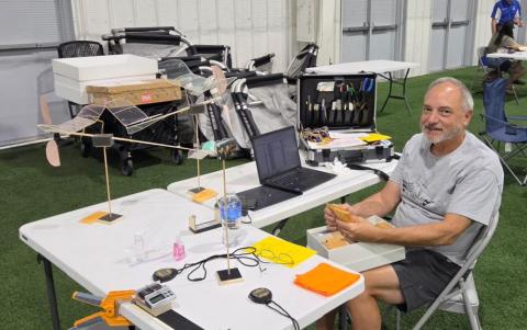 Man building model planes at indoor event, seated at a table with tools and laptop.