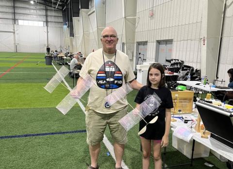 Man and girl holding model planes on indoor sports field.