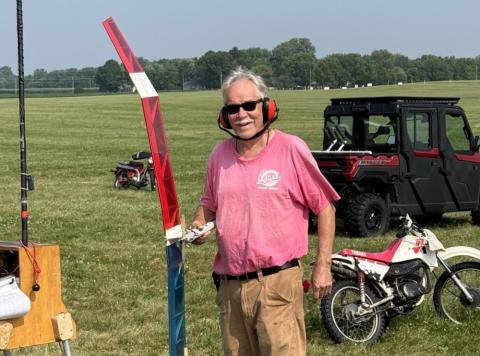 Man in pink shirt outdoors, wearing ear protection, near a red pole and motorcycles.